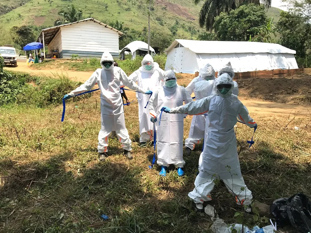 Operatori sanitari in tuta di protezione bianca disinfettano il terreno in una zona rurale con case di lamiera sullo sfondo.