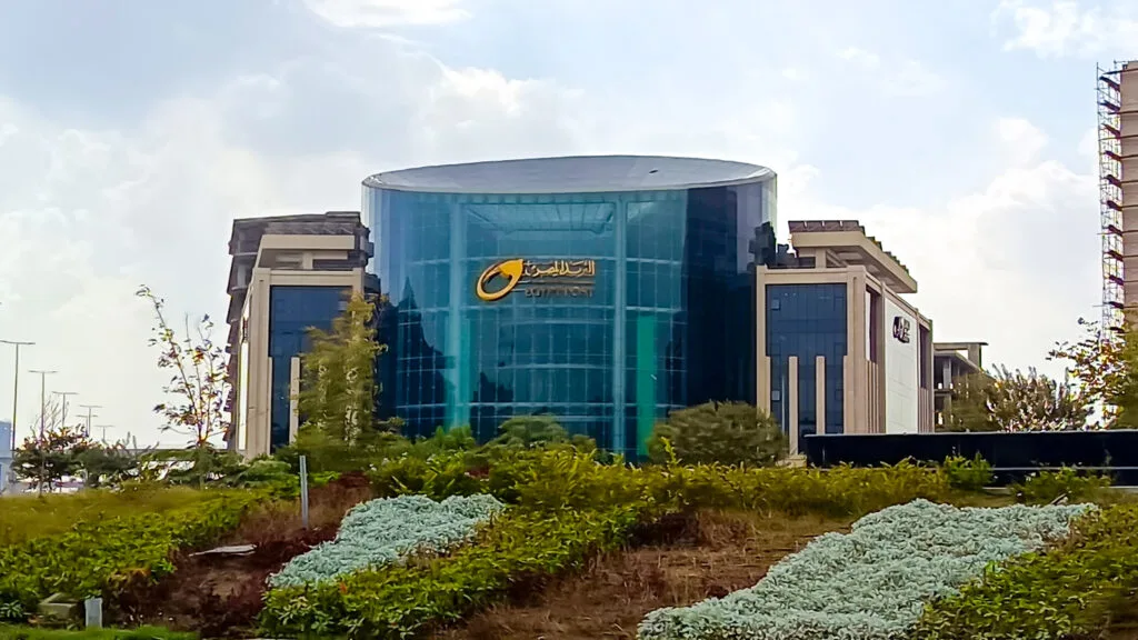 Edificio cilindrico in vetro blu con logo egiziano, circondato da vegetazione e strutture in laterizio nel deserto.