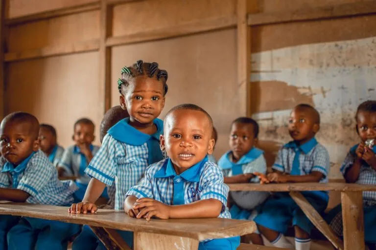 Bambini sorridenti in uniforme scolastica blu seduti su panche di legno in un'aula con pareti spoglie.