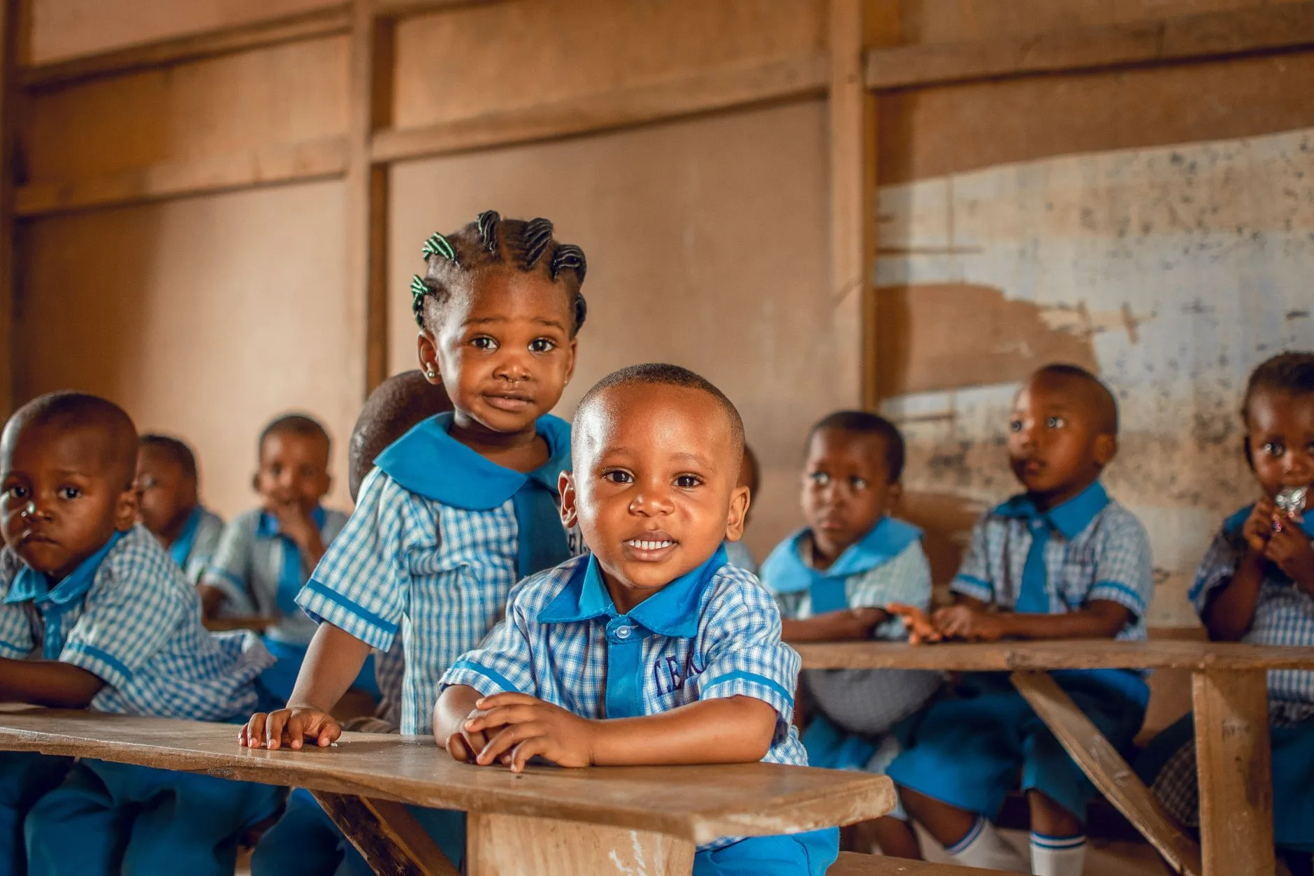Bambini sorridenti in uniforme scolastica blu seduti su panche di legno in un'aula con pareti spoglie.