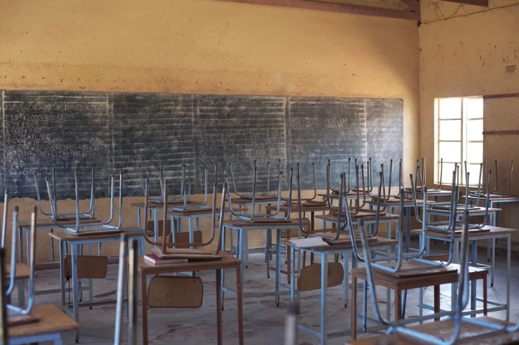 Aula scolastica vuota con banchi rovesciati su sedie e lavagna nera sullo sfondo, pareti ingiallite dalla polvere.
