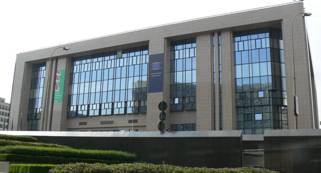 Edificio istituzionale moderno con ampia vetrata e bandiere, circondato da verde.