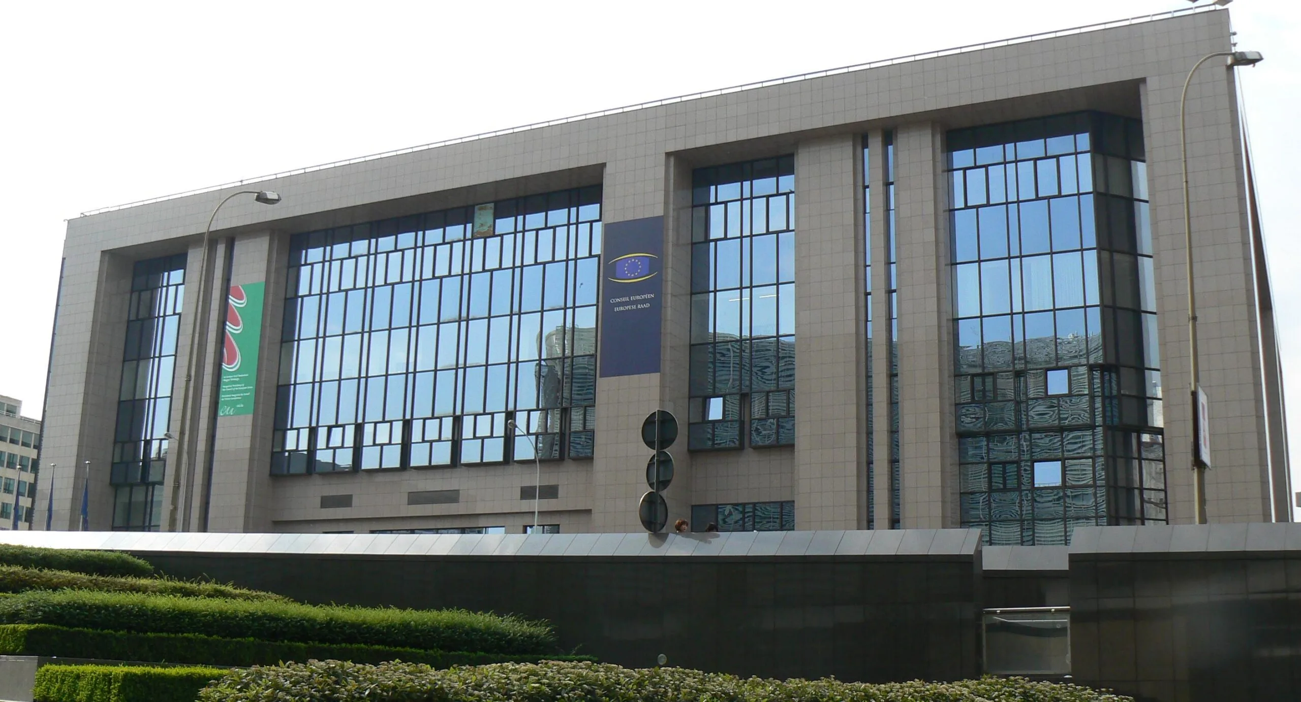 Edificio istituzionale moderno con ampia vetrata e bandiere, circondato da verde.