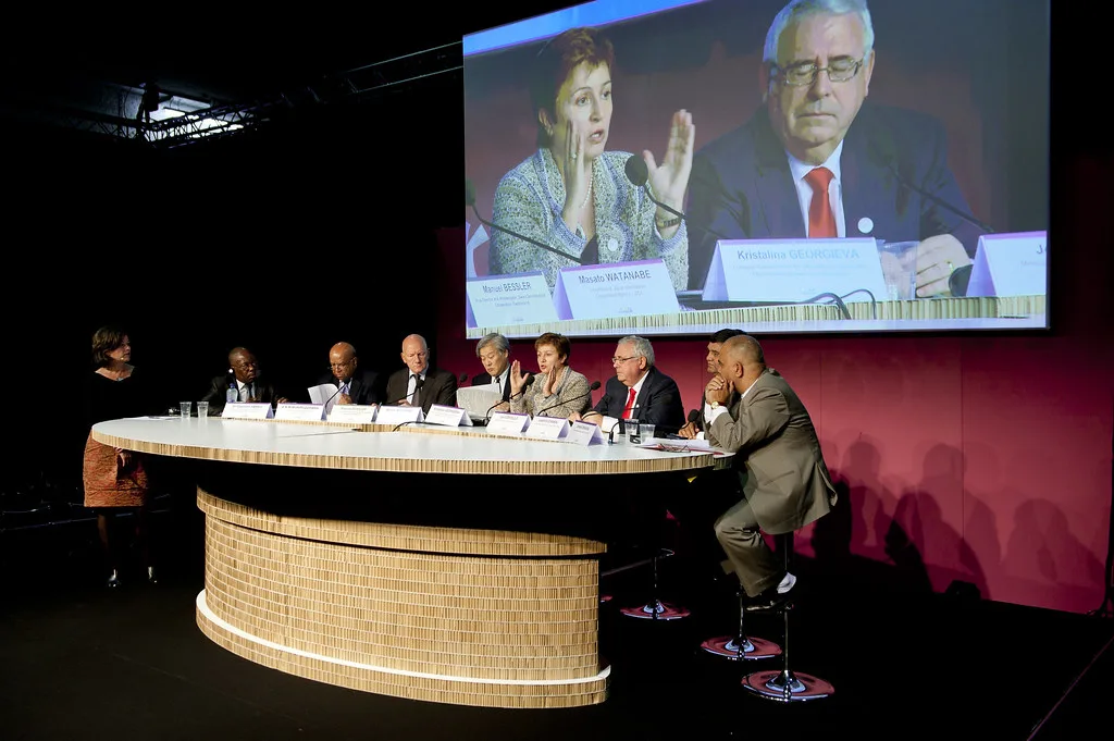 Tavolo rotondo con panel di discussione in auditorium; grande schermo retrostante mostra due relatori in primo piano.