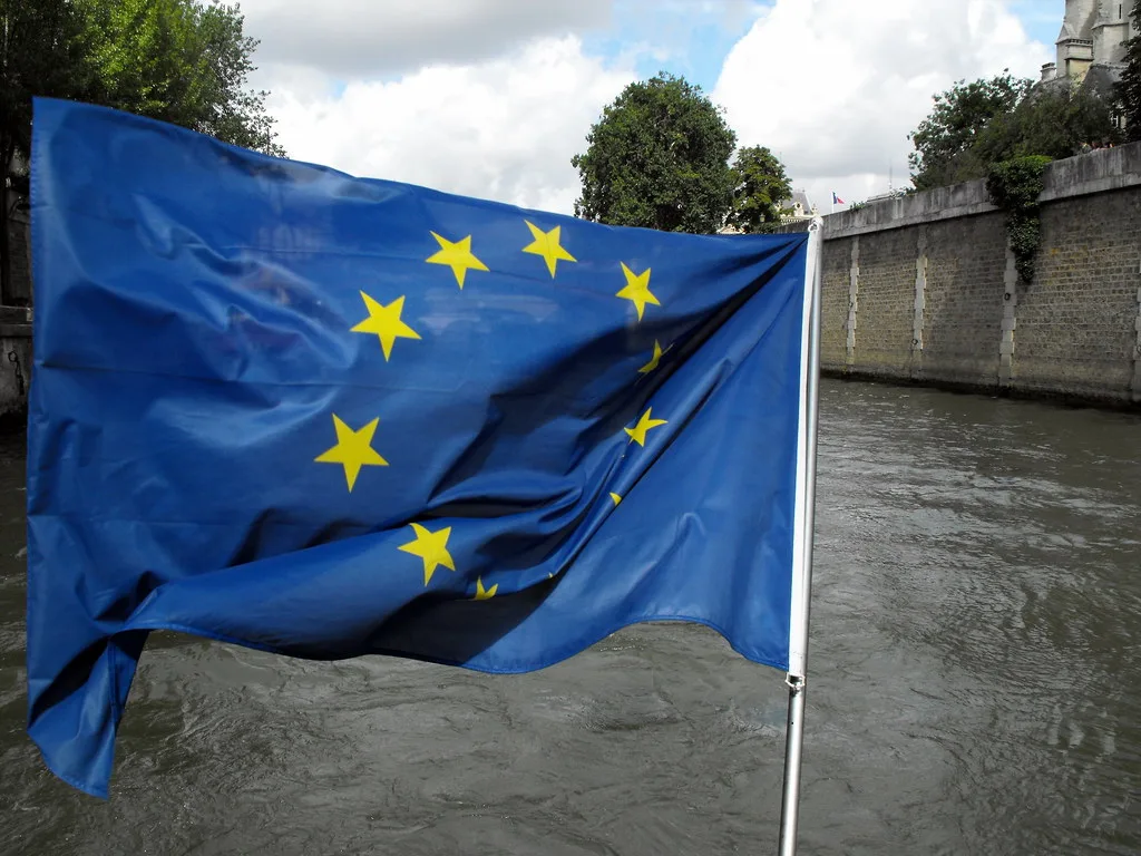 Bandiera dell'Unione Europea sventola davanti a un canale, con edifici storici sullo sfondo.