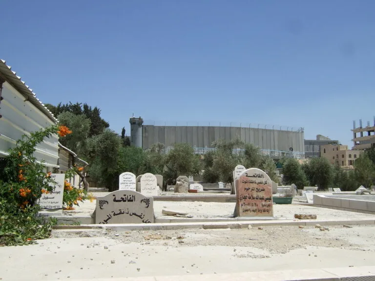 Cimitero con lapidi in arabo in primo piano, muro di cemento grigio e case in pietra sullo sfondo sotto cielo sereno.