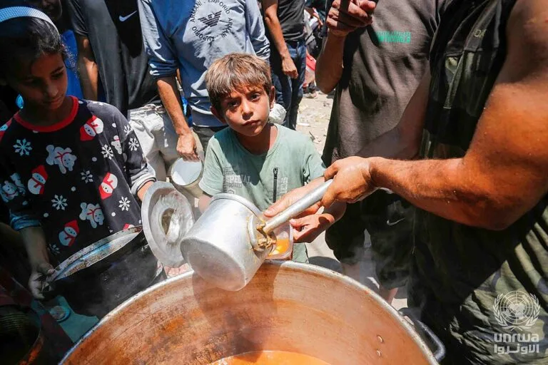 Bambini e adulti attorno a una pentola ricevono cibo in ciotole durante una distribuzione di aiuti.