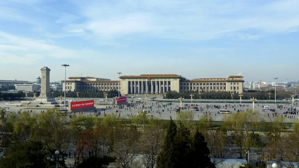 Piazza urbana con edificio monumentale in stile sovietico, monumento commemorativo e folla di persone sparse.