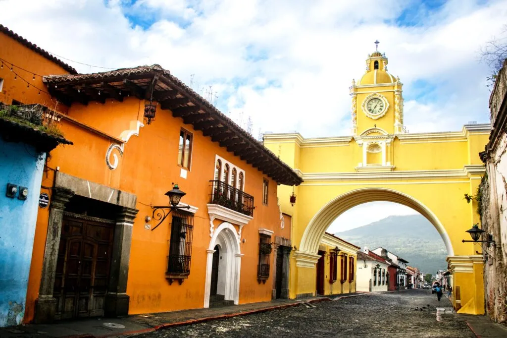 Strada coloniale con edifici colorati e arco monumentale giallo con torre dell'orologio, montagne sullo sfondo.