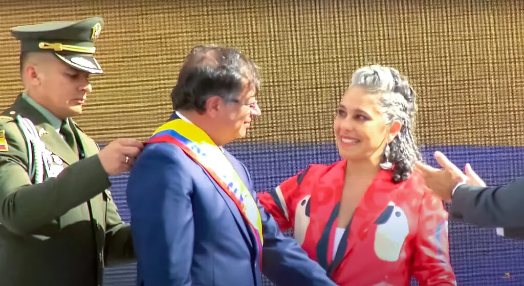 Ufficiale militare e donna in rosso accanto a uomo con fascia presidenziale colombiana durante cerimonia ufficiale.