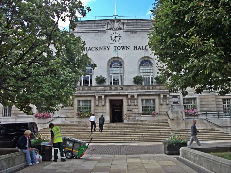Hackney Town Hall con facciata in pietra grigia, orologio e iscrizione, circondata da alberi e persone in piazza.