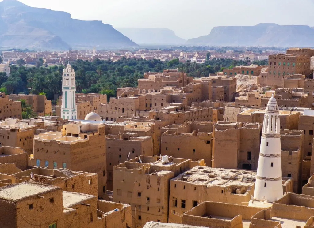 Città yemenita con edifici in mattoni di fango, minareti bianchi e montagne desertiche sullo sfondo.