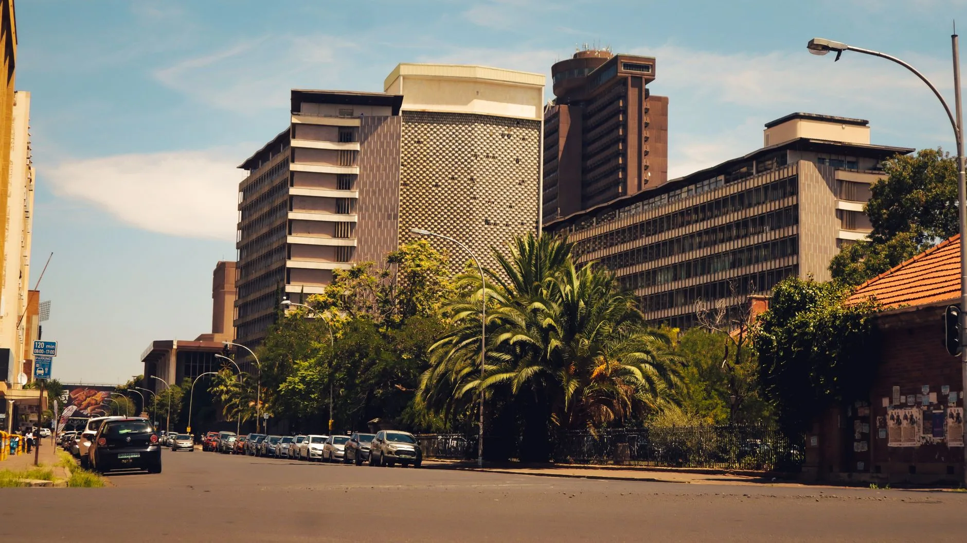 Strada urbana con edifici moderni di diversi piani, palme e auto parcheggiate sotto un cielo sereno.