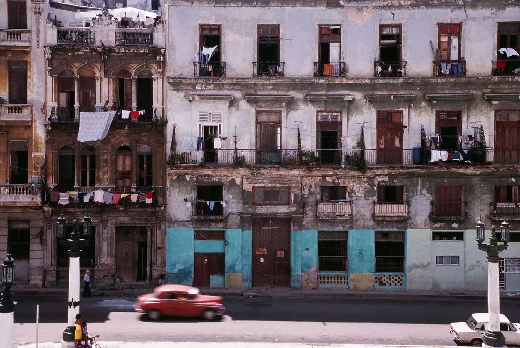Palazzo coloniale deteriorato all'Avana con facciate sgualcite, finestre scure e bucato ai balconi; un'auto rossa passa…