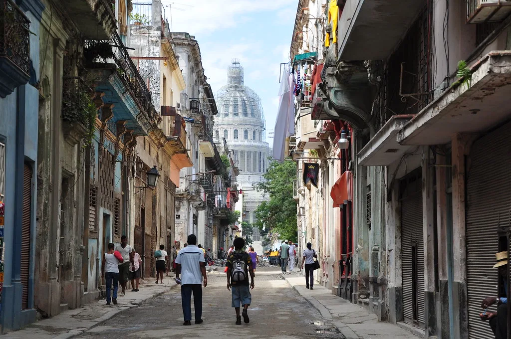 Strada dell'Avana con edifici coloniali logori e la cupola del Campidoglio visibile sullo sfondo tra i passanti.