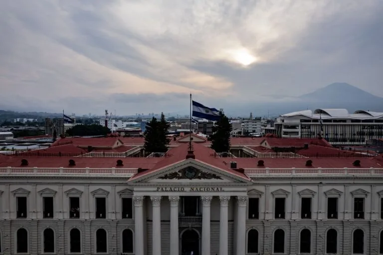 Palazzo Nazionale con colonnato bianco e tetto rosso, bandiera sulla terrazza e vulcani sullo sfondo.