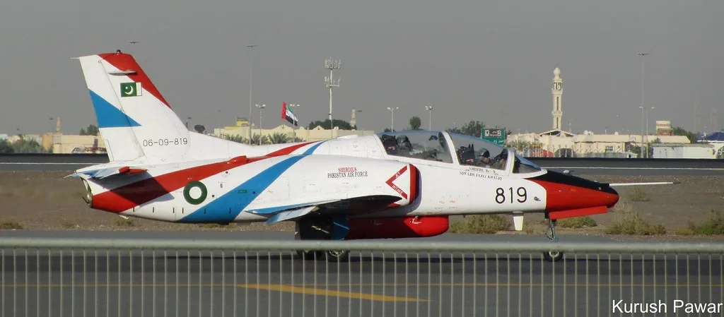 Aereo da addestramento bianco e rosso con numero 819 in pista aeroportuale; sullo sfondo moschea e skyline urbano.