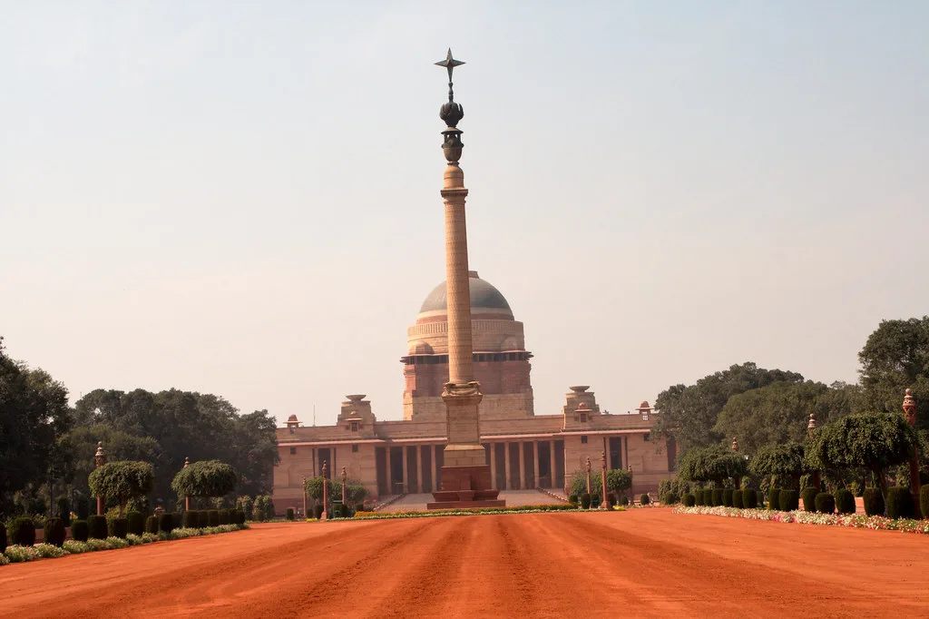 Edificio neoclassico con cupola e colonna centrale su un viale in terra battuta rossa, circondato da alberi in India.