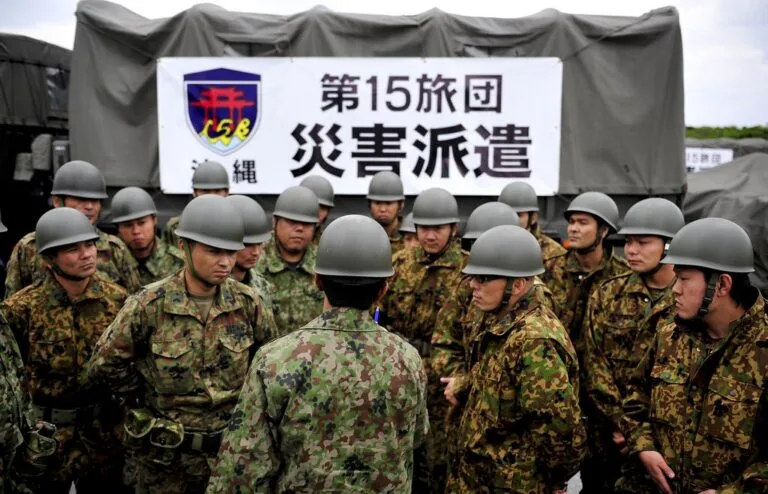 Militari giapponesi in divisa mimetica e elmetti davanti a un tendone con caratteri giapponesi e stemma dell'esercito.