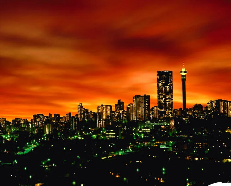 Lo skyline di Johannesburg illuminato di notte contro un cielo arancione, con grattacieli e luci verdi sparse.