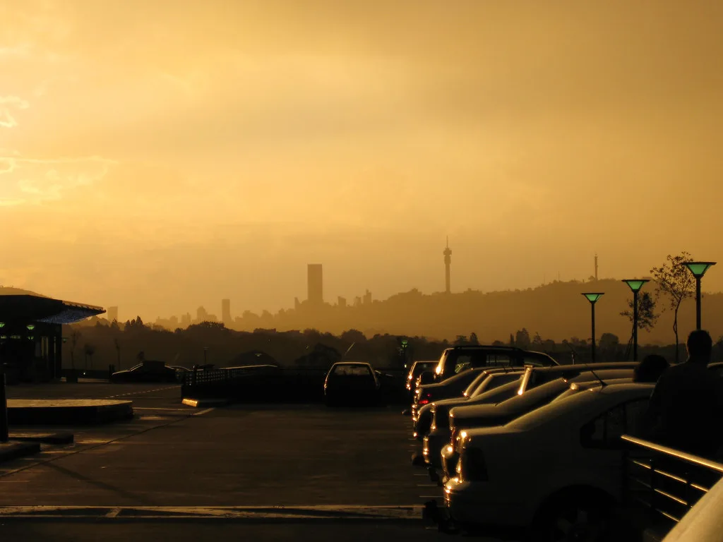 Skyline di Johannesburg avvolta da smog al tramonto, con edifici sfocati e luci artificiali in primo piano.