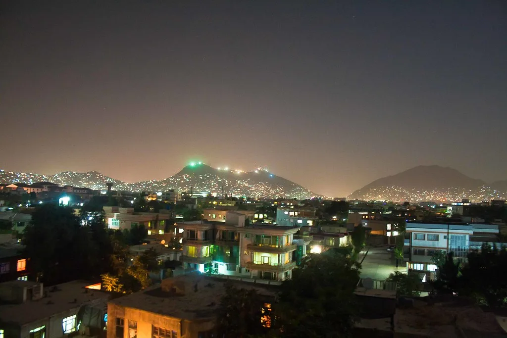 Veduta notturna di Kabul con edifici residenziali illuminati e montagne sullo sfondo coperte di luci.