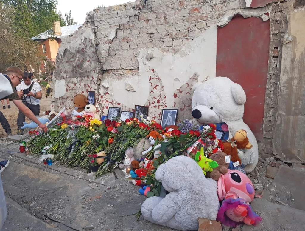 Muro danneggiato con foto, fiori e peluche in ricordo davanti a una porta rossa, persone in visita al monumento improvvisato.