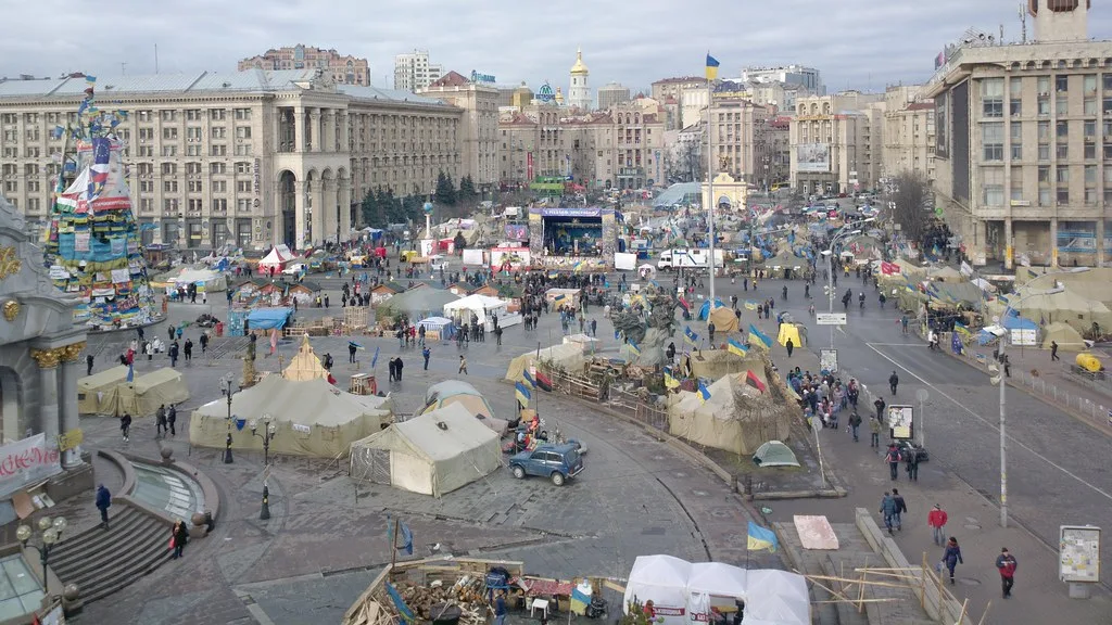Piazza centrale di Kiev con folla di manifestanti, edifici storici e bandiera ucraina in sfondo.