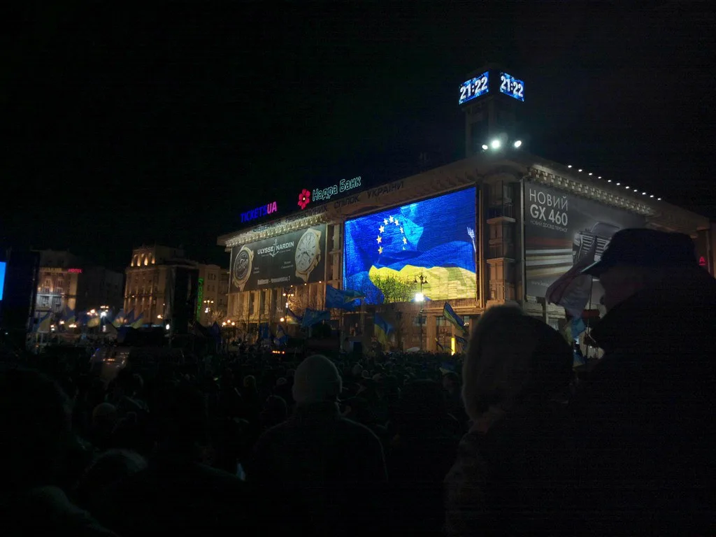 Folla radunata di notte in piazza davanti a grande schermo con colori azzurri e gialli, edifici storici illuminati sullo…
