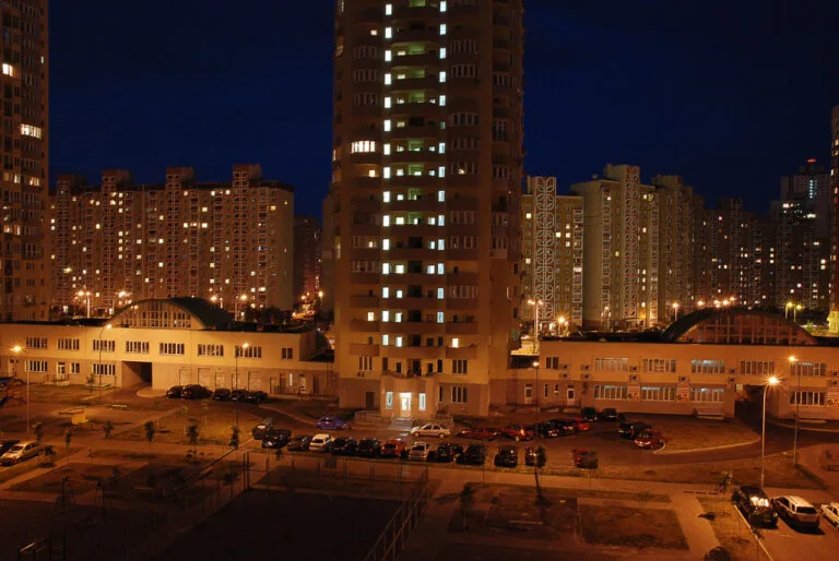 Complesso residenziale notturno con edifici illuminati, cortile interno con auto parcheggiate e cielo scuro.