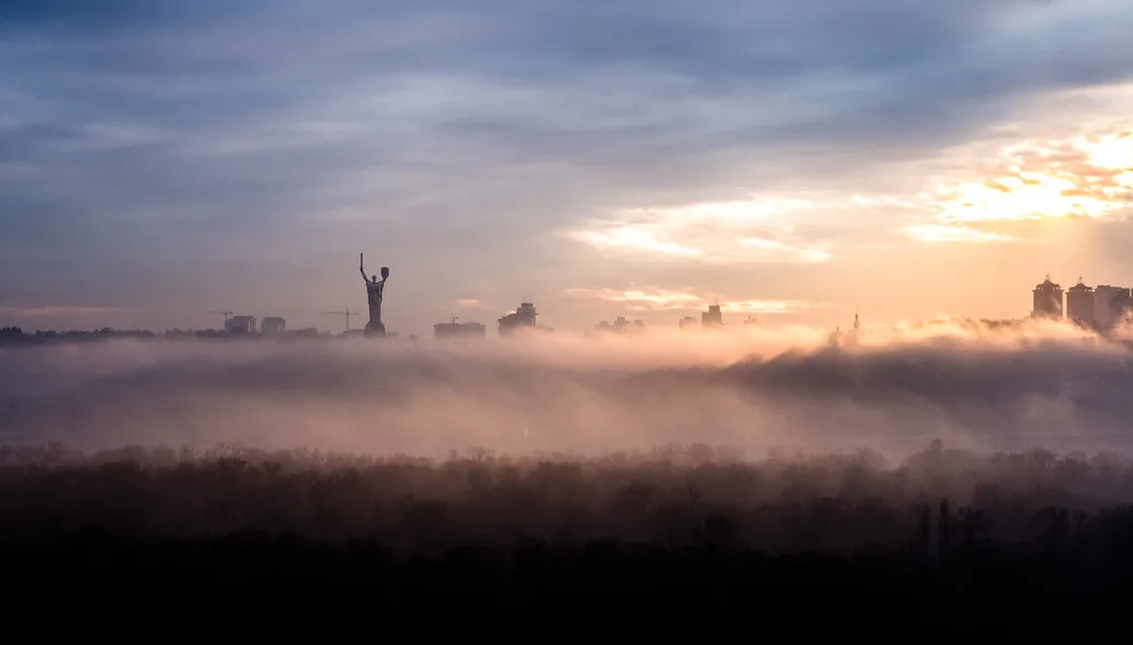Skyline di una città avvolta nella nebbia con una statua monumentale a braccia alzate e grattacieli sullo sfondo.