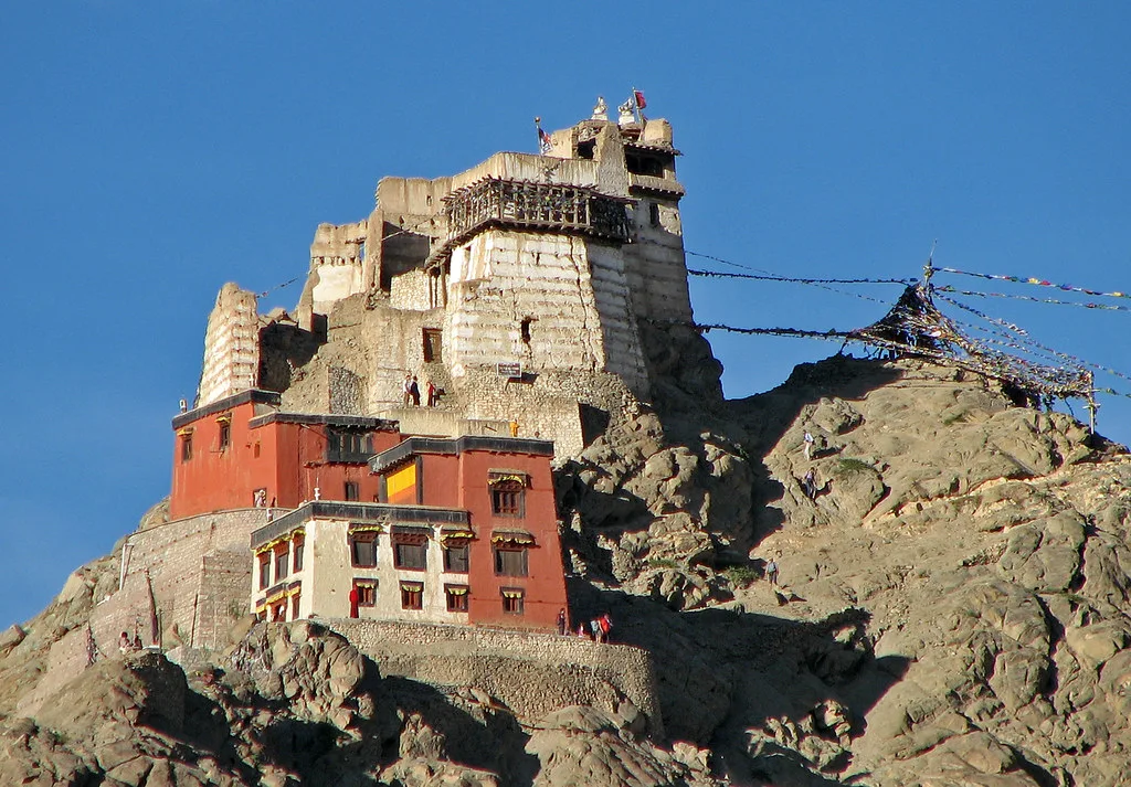 Palazzo antico arroccato su una roccia scura con strutture in pietra dorata sopra e una costruzione rossa in basso.