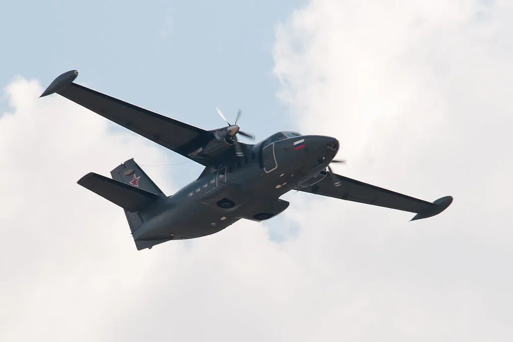 Aereo da trasporto militare nero a doppia elica fotografato in volo con cielo nuvolato sullo sfondo.