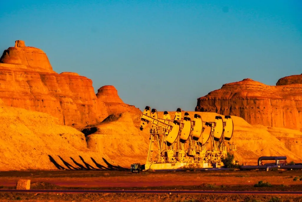 Pompe di estrazione petrolifera gialle allineate in un paesaggio desertico con formazioni rocciose arancioni al tramonto.
