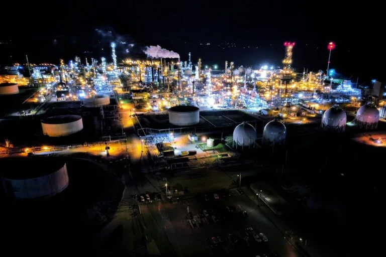 Complesso petrolifero illuminato di notte con serbatoi, torce di fiamma e strutture di raffinazione.