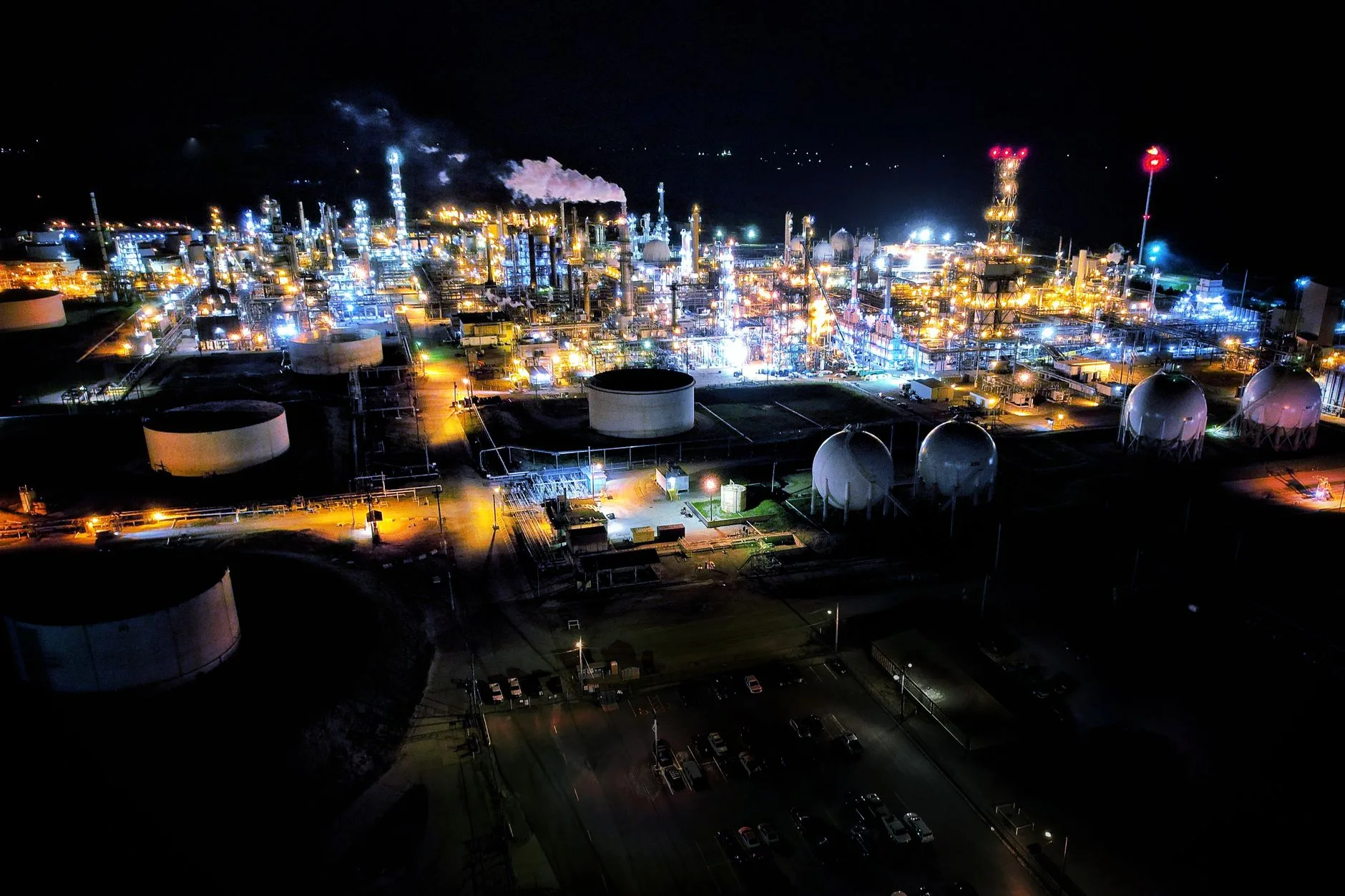 Complesso petrolifero illuminato di notte con serbatoi, torce di fiamma e strutture di raffinazione.