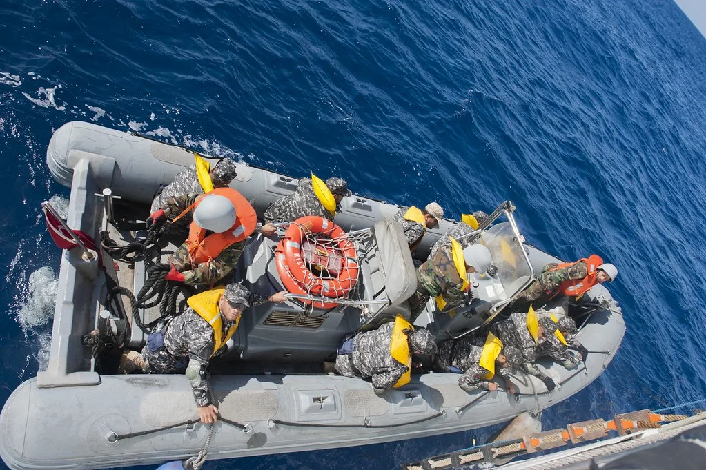 Operazione marittima con gommone di soccorso carico di persone in giubbotti arancioni su acque blu profonde.