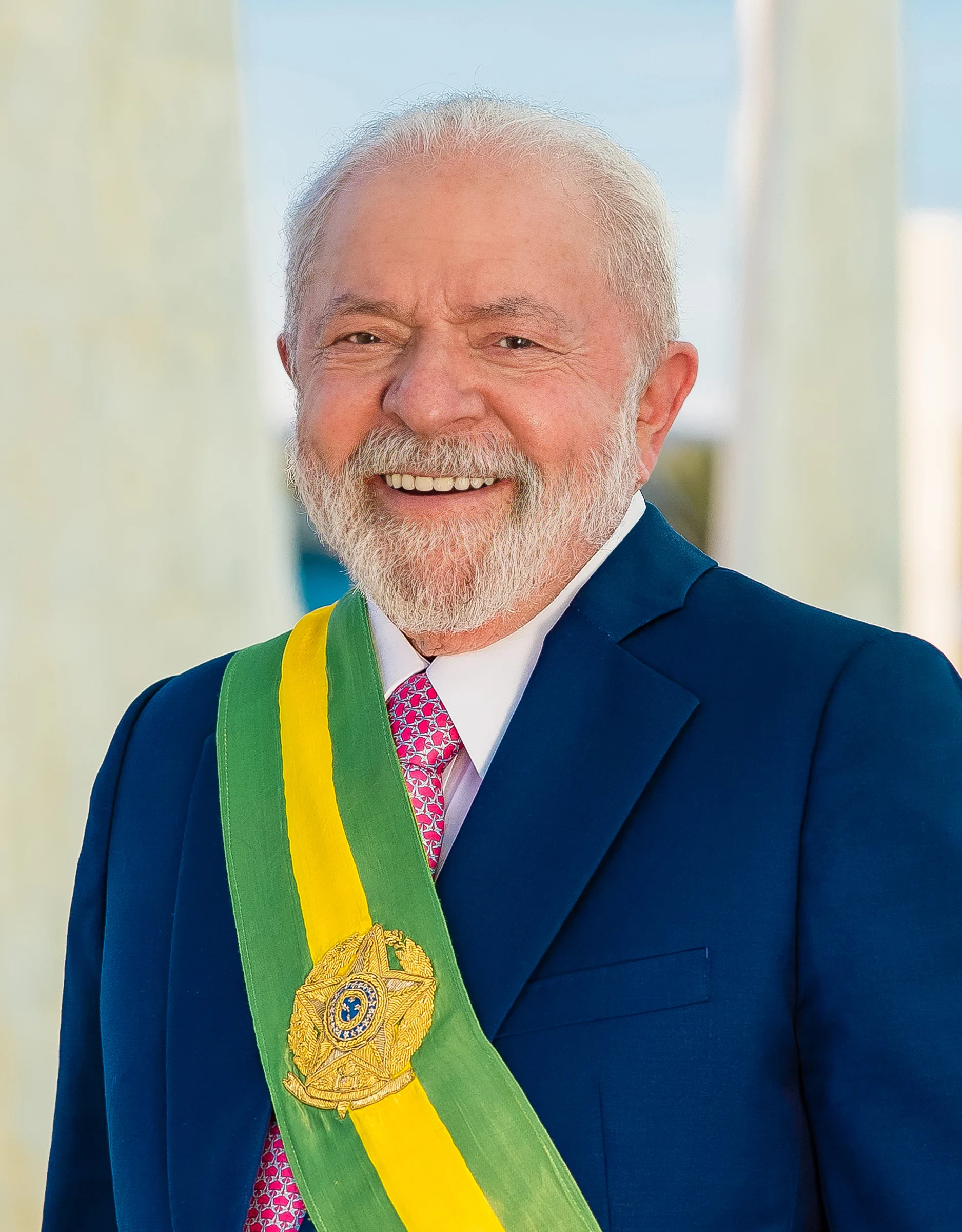 Uomo anziano con barba grigia in giacca blu e fascia presidenziale verde-gialla sorride verso la camera.
