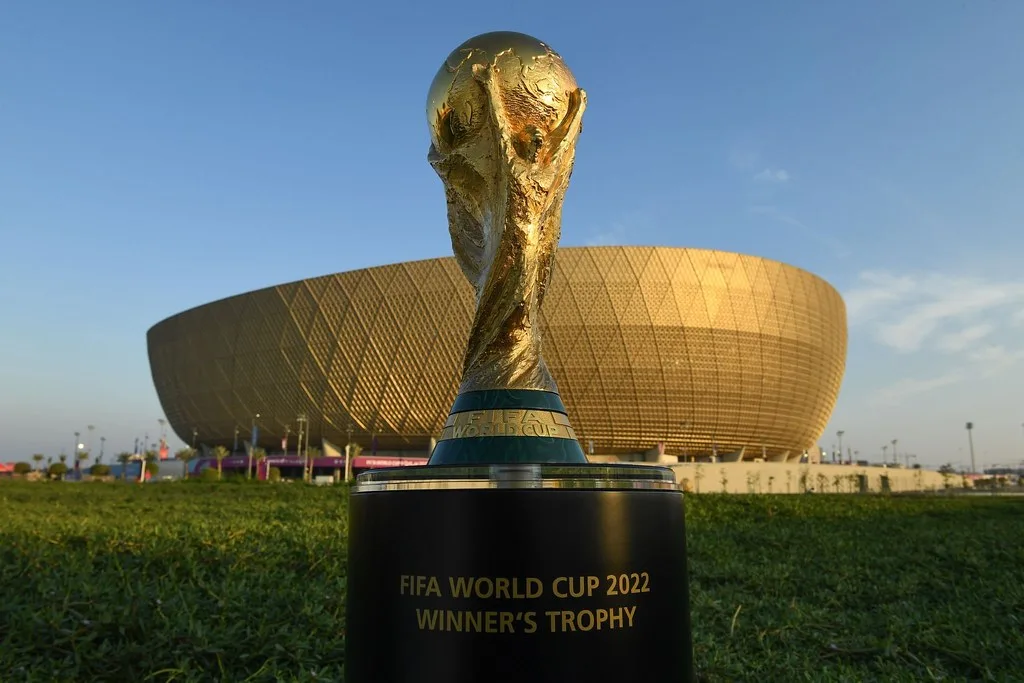 Trofeo della Coppa del Mondo FIFA dorato su piedistallo nero davanti allo stadio di Lusail con cielo sereno.