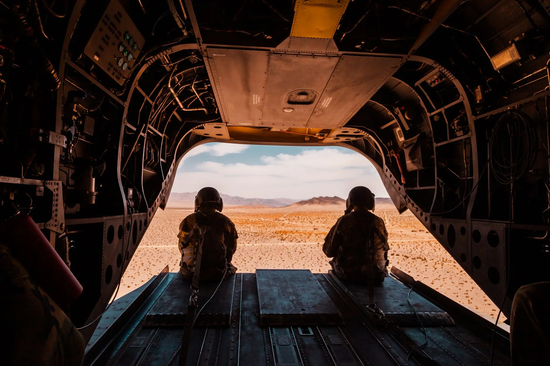 Due soldati seduti nel vano di carico di un elicottero militare osservano il vasto paesaggio desertico sottostante.