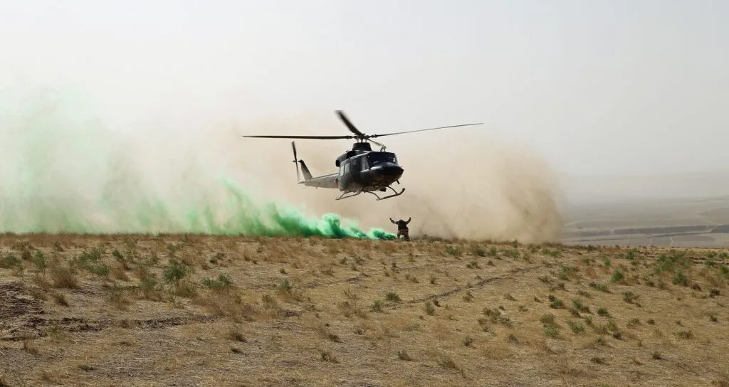 Elicottero militare in avvicinamento a un terreno desertico con fumo verde di segnalazione e soldato a terra.