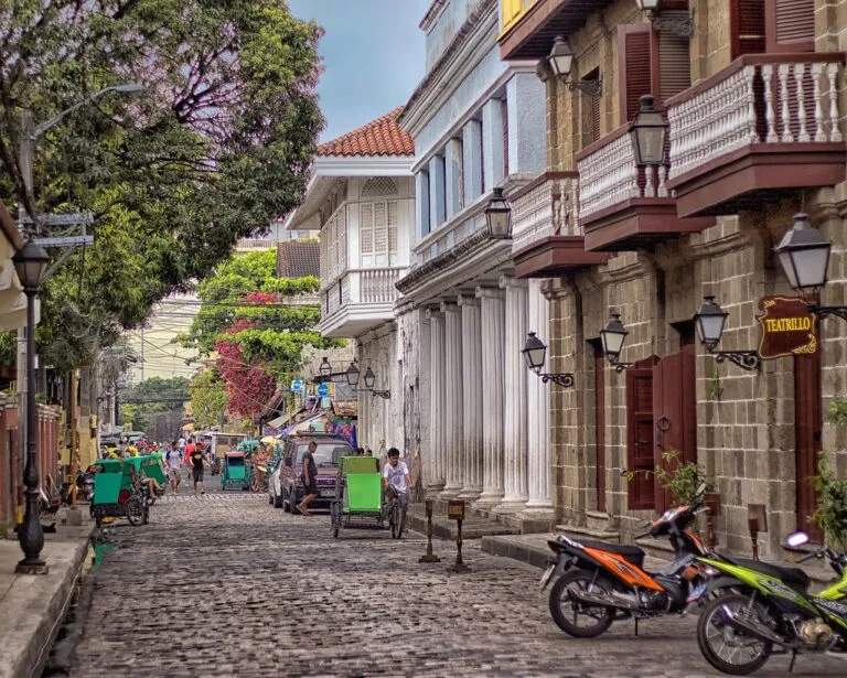 Strada acciottolata coloniale con edifici in stile spagnolo, balconate e carrozze tradizionali verdi.