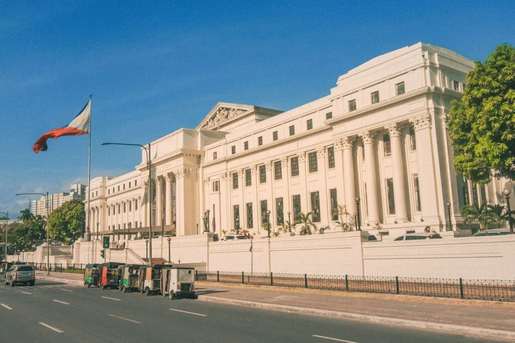 Palazzo neoclassico color crema con colonnati, bandiera filippina a sinistra, strada trafficata davanti e cielo sereno.