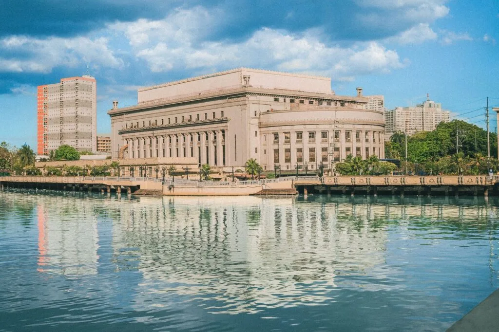 Palazzo neoclassico in pietra chiara affacciato su un fiume con riflesso, circondato da edifici moderni sullo sfondo.