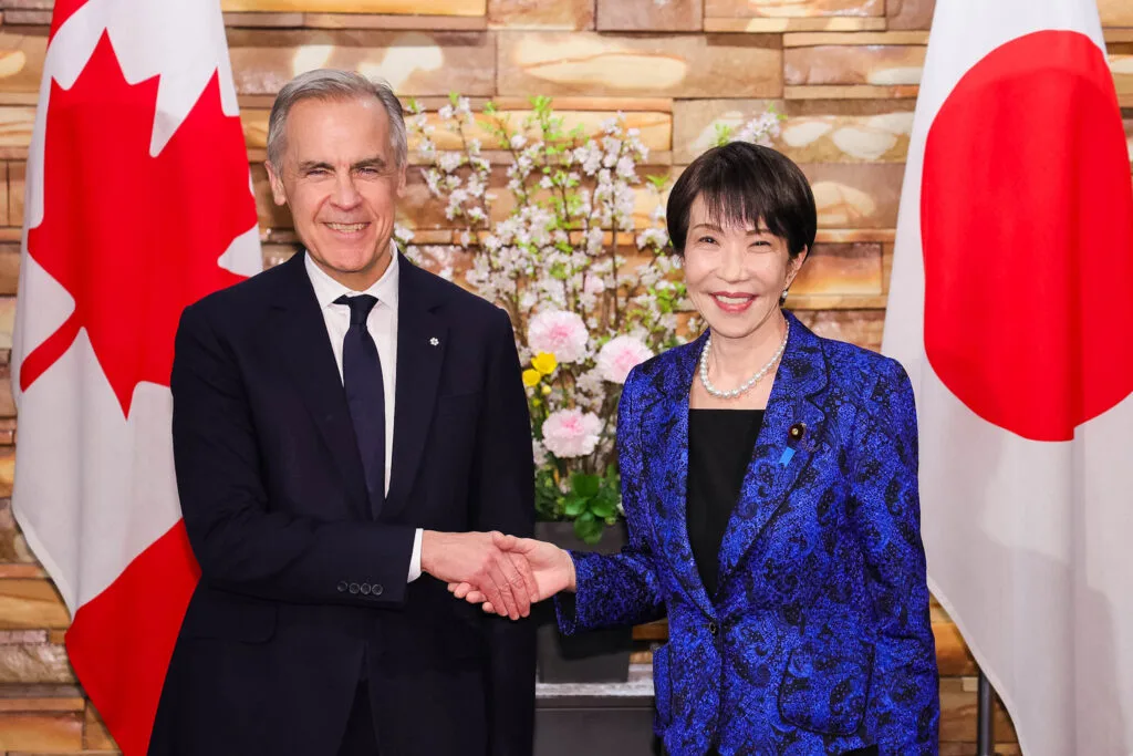 Due funzionari si stringono la mano davanti alle bandiere del Canada e del Giappone in una sala in pietra.