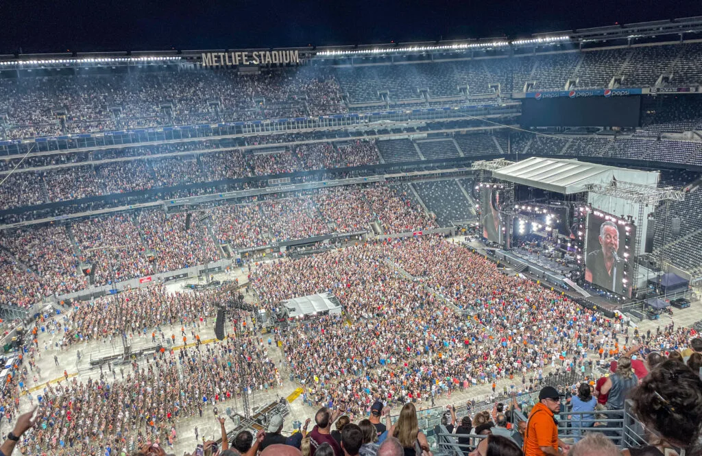 Stadio MetLife pieno di spettatori durante un grande evento dal vivo, con palco e schermi illuminati.