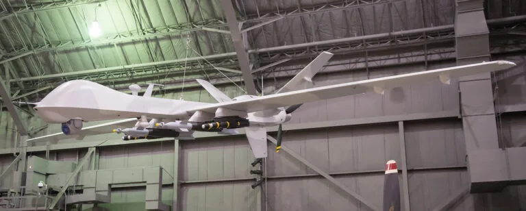 Drone militare grigio in hangar con carico di munizioni sottostanti, sospeso su strutture metalliche di officina.