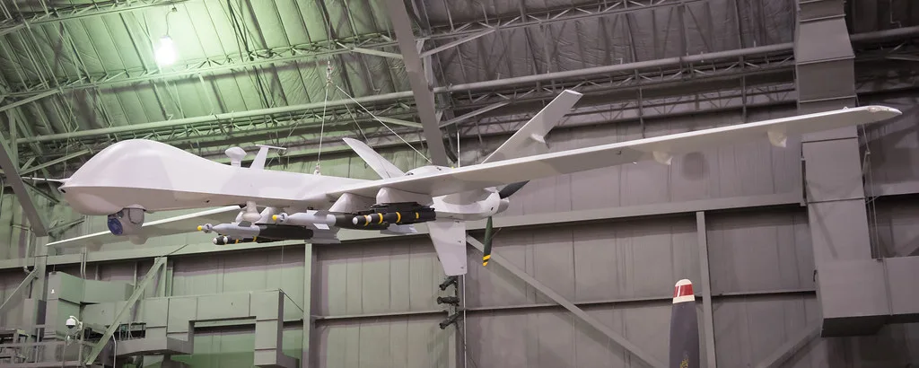 Drone militare grigio in hangar con carico di munizioni sottostanti, sospeso su strutture metalliche di officina.