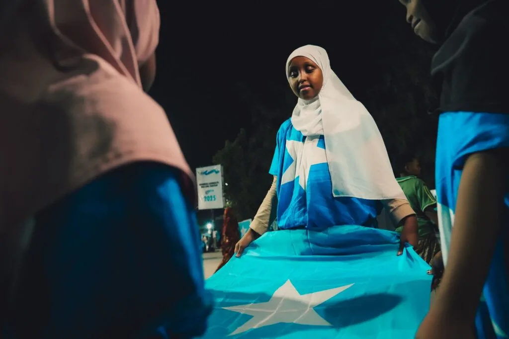 Giovane donna con hijab bianco e giacca blu regge la bandiera somala durante una celebrazione notturna.