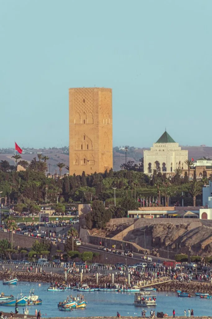 Torre Hassan e Mausoleo di Mohammed V a Rabat con bandiera marocchina, spiaggia affollata e barche nel porto sottostante.